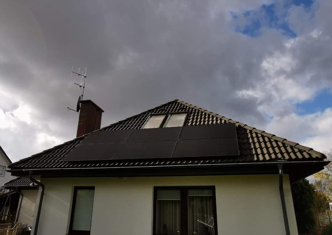 Ciemne panele słoneczne na brązowym dachu domu jednorodzinnego z kominem i anteną telewizyjną, na tle pochmurnego nieba.