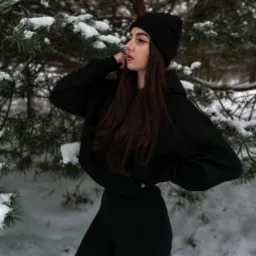 Kobieta w czarnym dresie i czapce pozuje na tle ośnieżonych gałęzi sosny, odwr&oacute;cona profilem w lewo, z dłonią uniesioną do szyi.