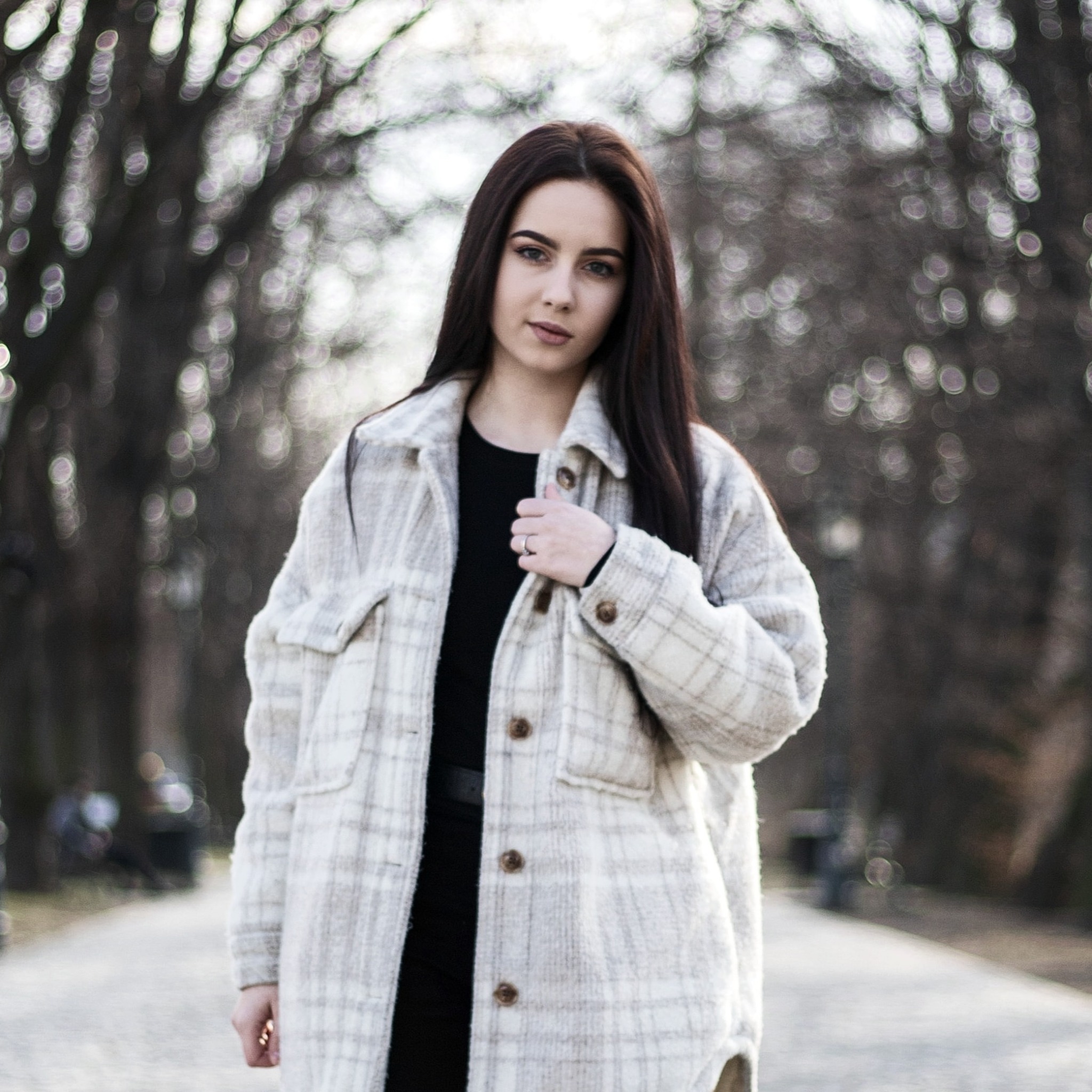 Portret młodej kobiety z długimi, ciemnymi włosami, w jasnej kraciastej kurtce na tle rozmytej alei parkowej z bokeh.