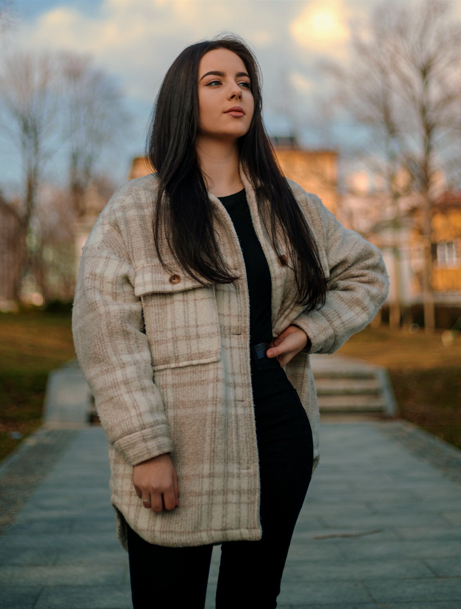 Portret młodej kobiety z długimi, ciemnymi włosami, ubranej w czarny top i beżową, kraciastą kurtkę oversize, pozującej na tle parku z budynkami w oddali.