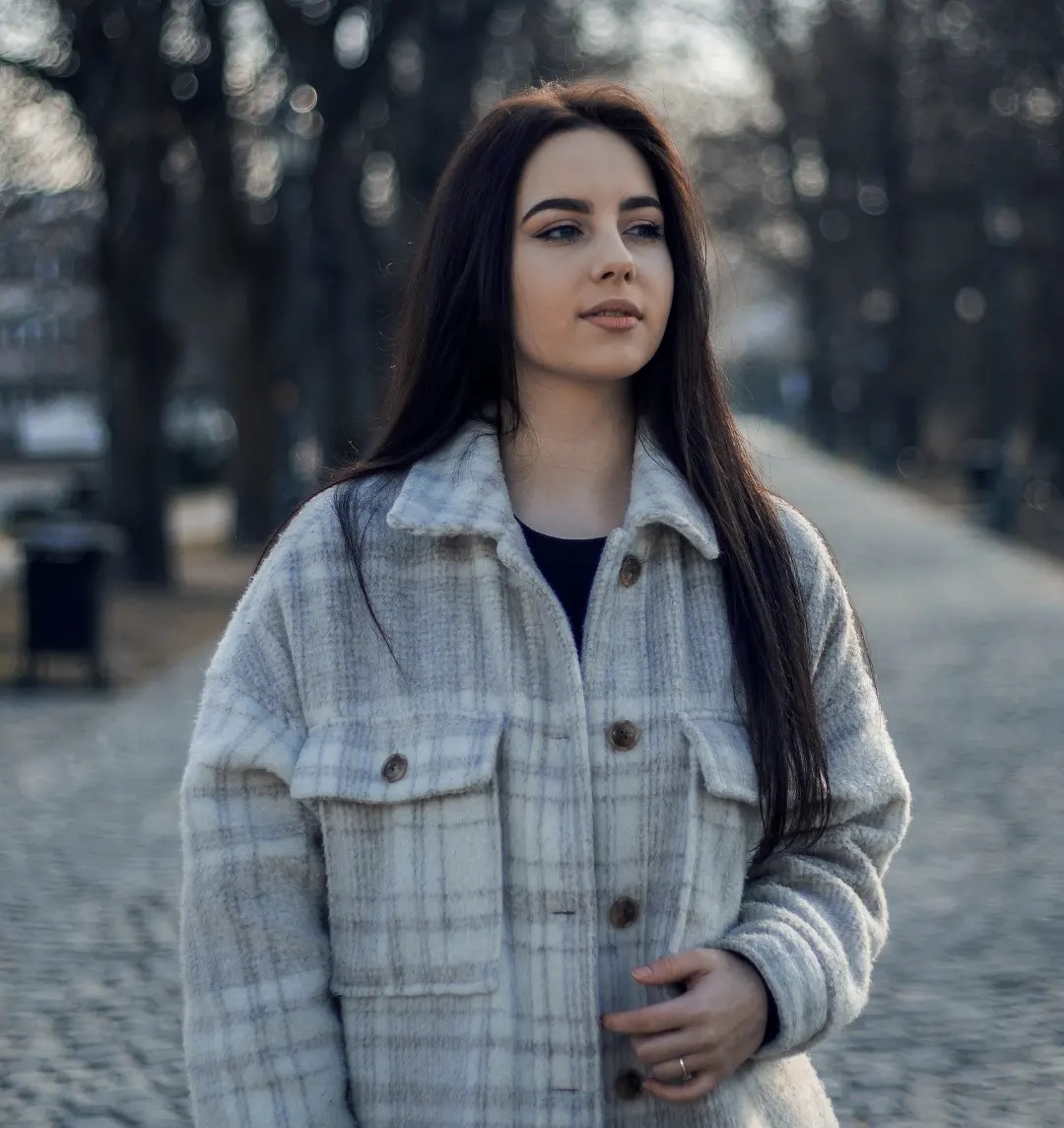 Portret młodej kobiety z długimi, ciemnymi włosami w jasnej, flanelowej kurtce w kratę, stojącej na brukowanej ścieżce w parku, z zamyślonym wyrazem twarzy i delikatnym makijażem.