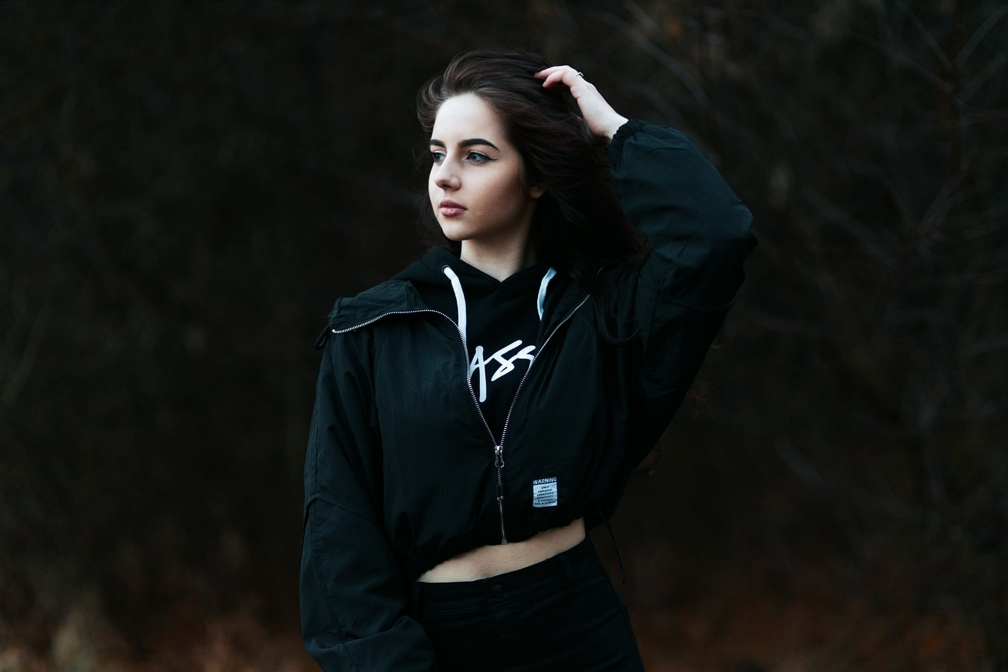Młoda kobieta z ciemnymi włosami, w czarnej kurtce i bluzie z kapturem, pozuje na tle ciemnego, rozmytego tła, delikatnie dotykając włosów dłonią z pierścionkiem.