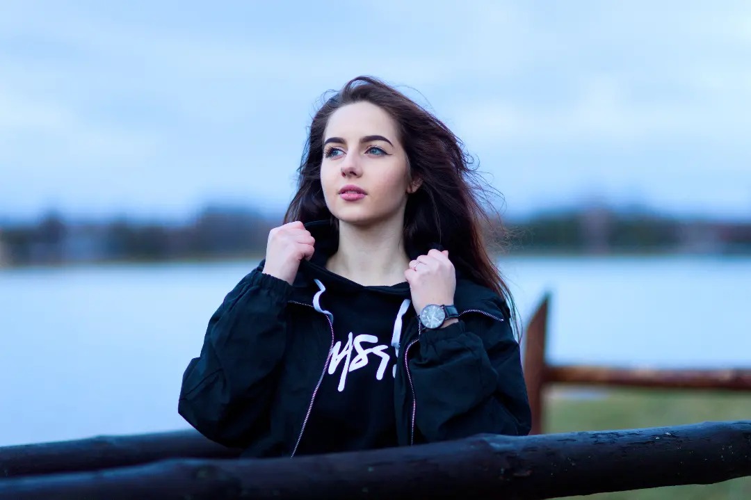 Portret młodej kobiety z długimi, brązowymi włosami, w czarnej bluzie z kapturem i kurtce, stojącej na tle jeziora i pochmurnego nieba, opierającej się o drewnianą barierkę i trzymającej dłonie...