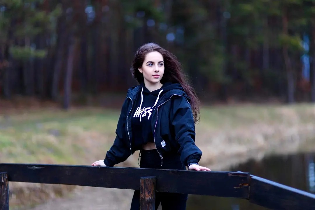 Portret młodej kobiety w czarnej kurtce i bluzie z kapturem, opierającej się o drewnianą barierkę mostku na tle zalesionego terenu i wody.