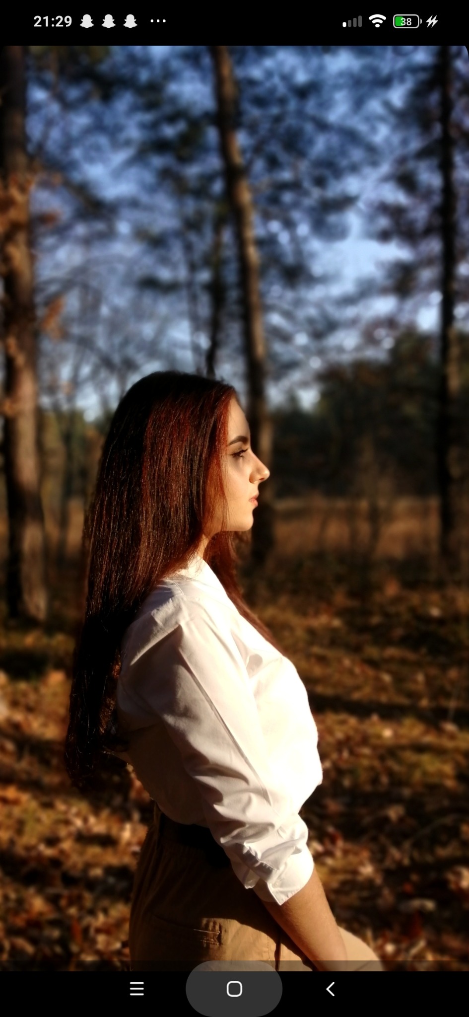 Portret kobiety z profilu na tle jesiennego lasu, oświetlona ciepłym światłem słonecznym, ubrana w białą koszulę i beżowe spodnie.