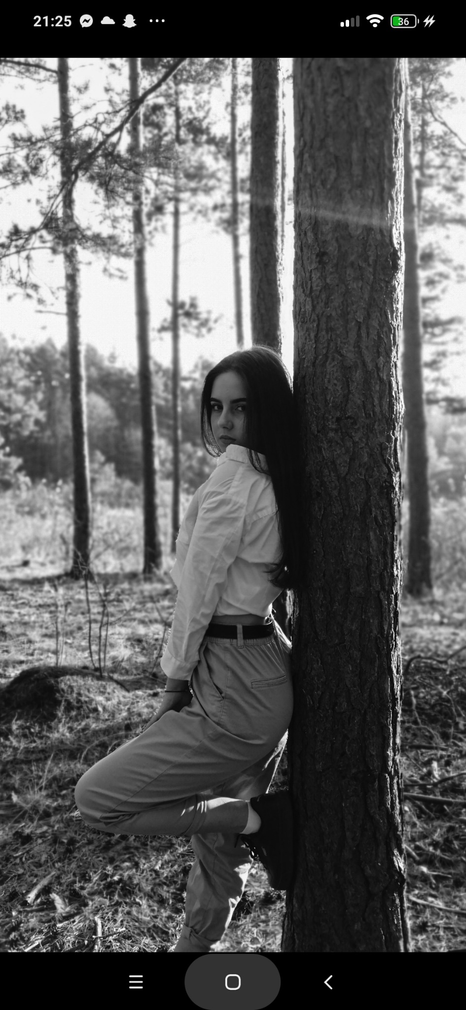 Czarno-białe zdjęcie kobiety opierającej się o sosnę w lesie, spoglądającej przez ramię. Ubrana w jasną koszulę i spodnie, z długimi, ciemnymi włosami.