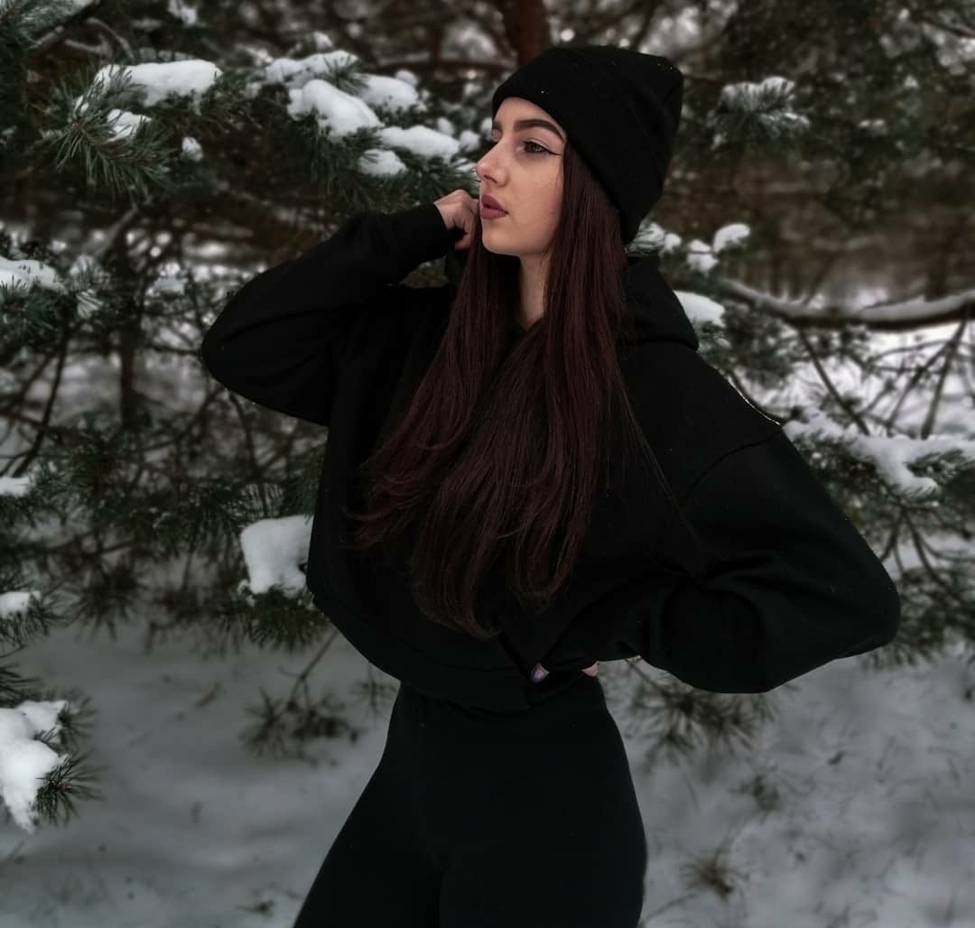 Kobieta w czarnym dresie i czapce pozuje na tle ośnieżonych gałęzi sosny, odwrócona profilem w lewo, z dłonią uniesioną do szyi.