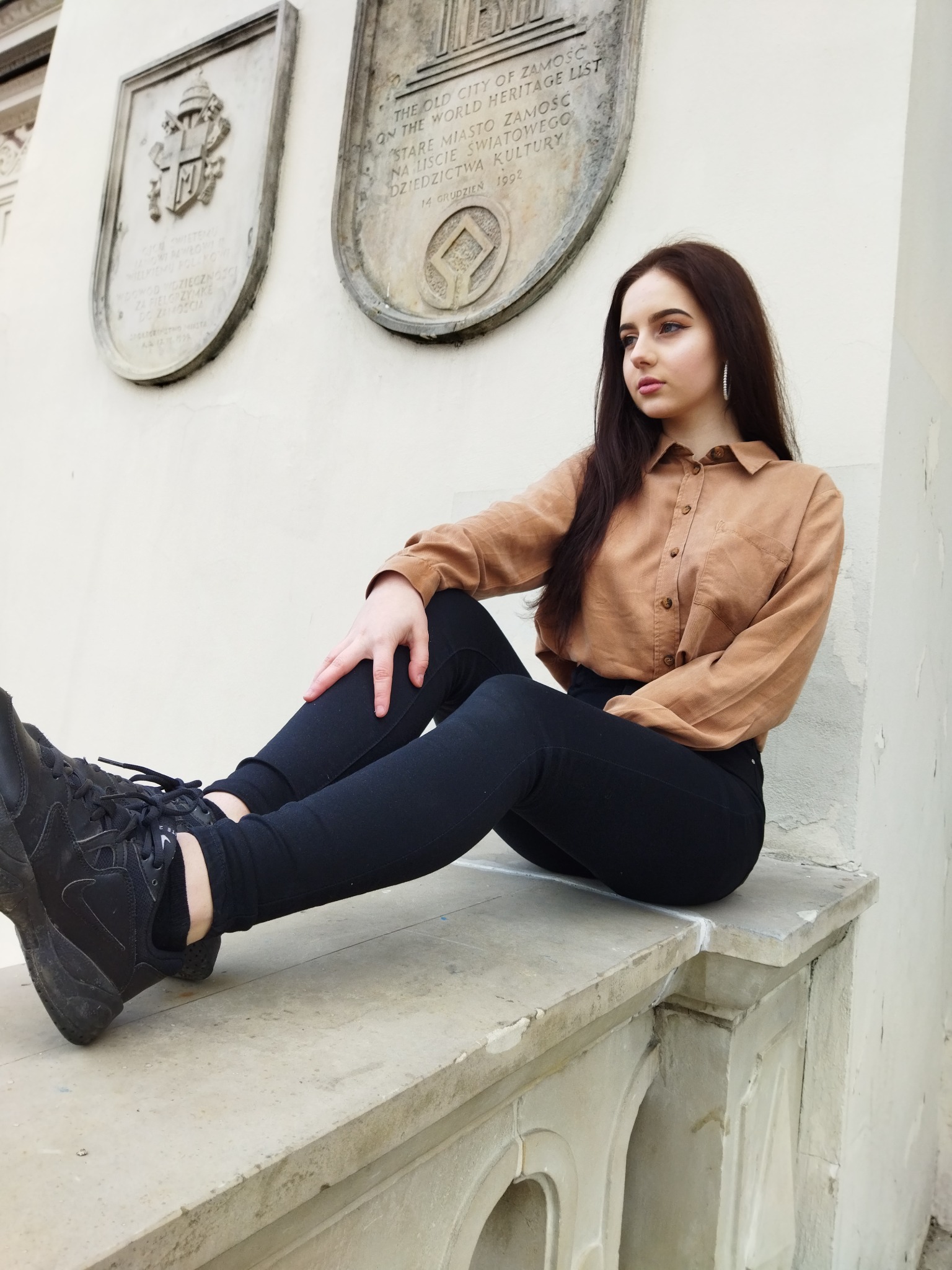 Portret młodej kobiety siedzącej na kamiennym murku w miejskim otoczeniu, w tle tablice pamiątkowe z inskrypcjami, ubrana w brązową koszulę i czarne spodnie.