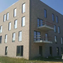 Nowoczesny budynek mieszkalny z jasnej cegły z balkonami z drewnianymi barierkami i dużymi oknami, widoczny samochód dostawczy z przodu.