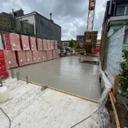 Świeżo wylany fundament z widocznymi przewodami, szalunkiem i paletami cegieł w tle, na budowie z dźwigiem w pochmurny dzień.