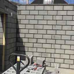 Wznoszenie ściany z szarych bloczków betonowych, widoczny fragment ściany, paleta z bloczkami, wiadro z zaprawą i narzędzia murarskie, na górze notyfikacja z Messengera.