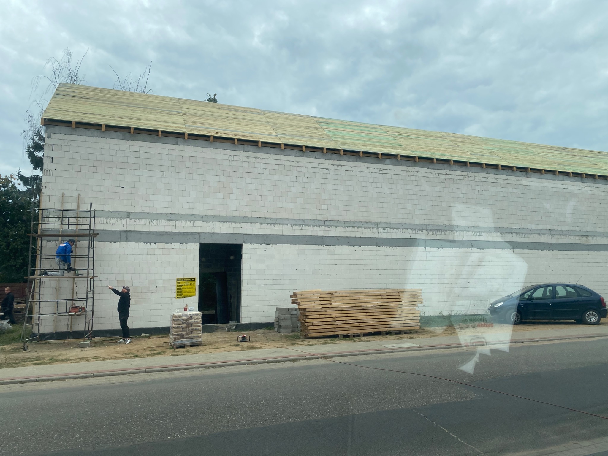 Wznoszenie dwukondygnacyjnego budynku z białych bloczków, widoczne rusztowanie z pracującym robotnikiem, stos desek i worki z zaprawą, na tle pochmurnego nieba i zaparkowanego samochodu.