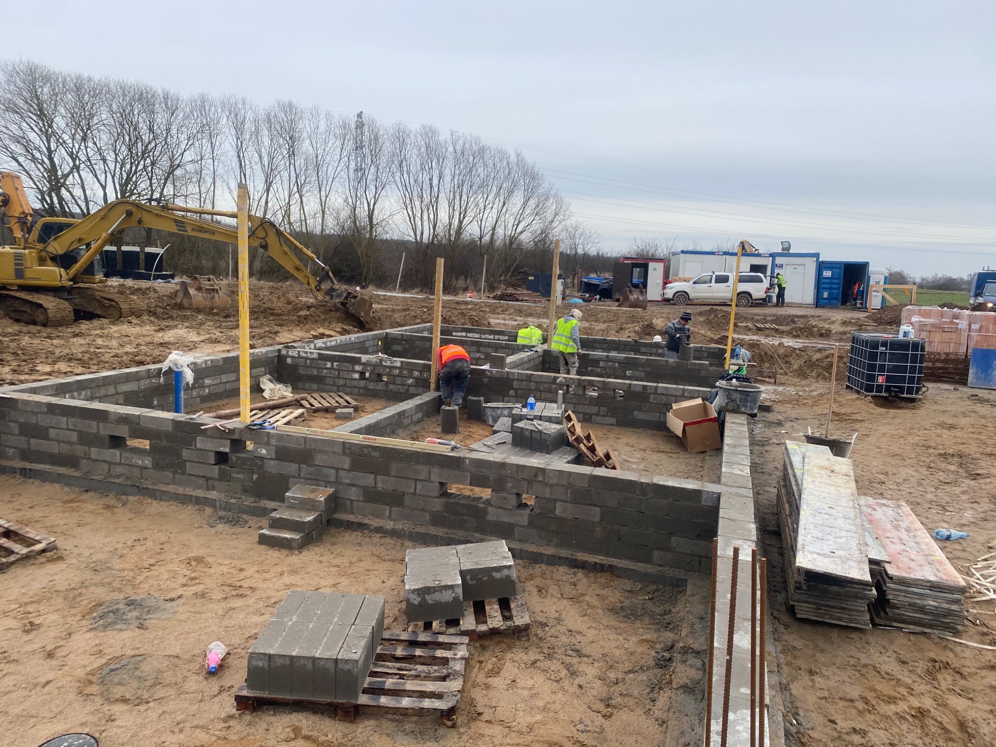 Budowa fundamentów z bloczków betonowych, widoczni pracownicy w odblaskowych kamizelkach, koparka w tle, teren budowy w trakcie prac.