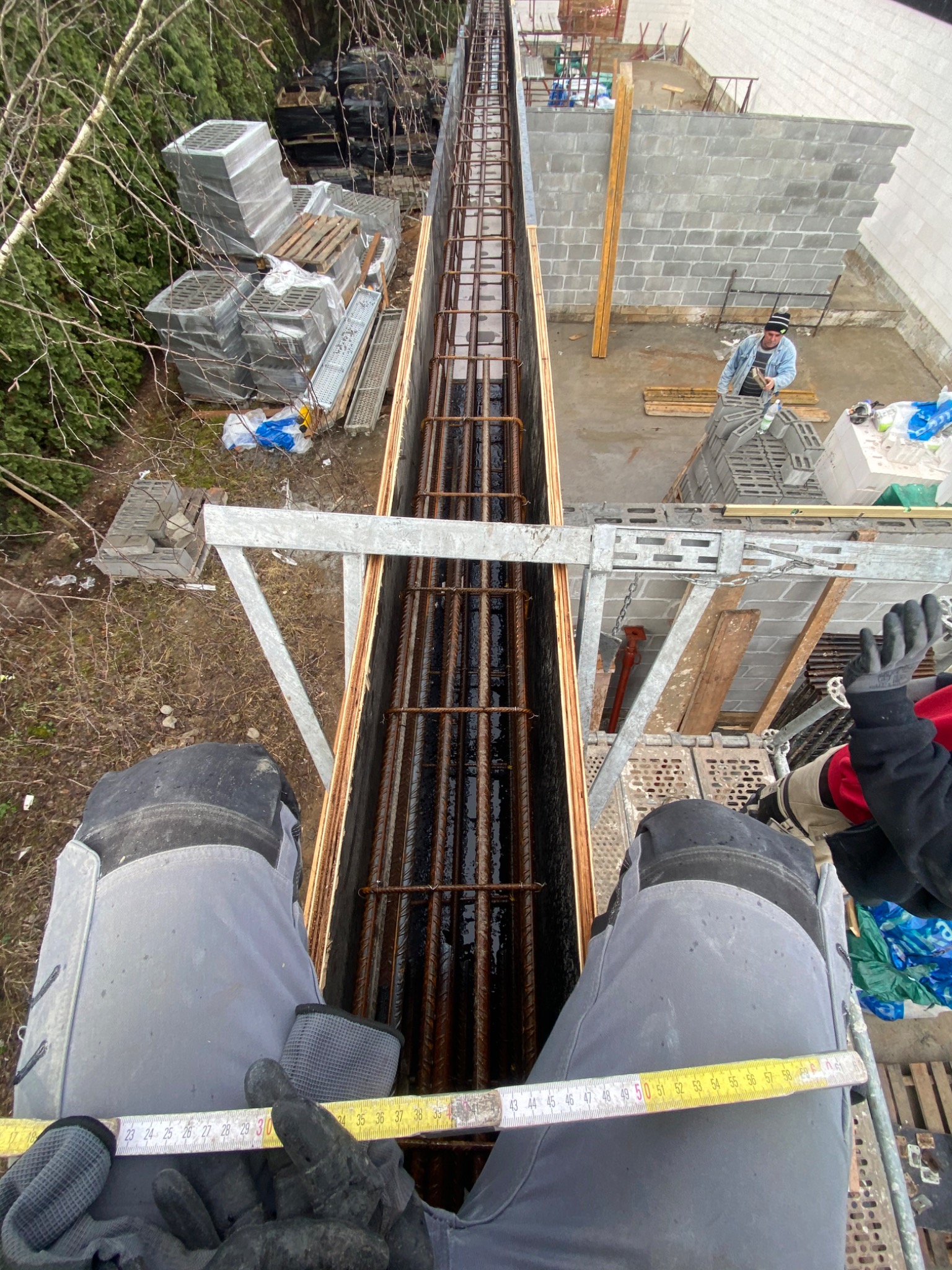 Widok z góry na przygotowany szalunek zbrojeniowy wypełniony stalowymi prętami, mierzony przez pracownika w roboczych spodniach i rękawicach, w tle mur z bloczków betonowych i pracujący mężczyzna.