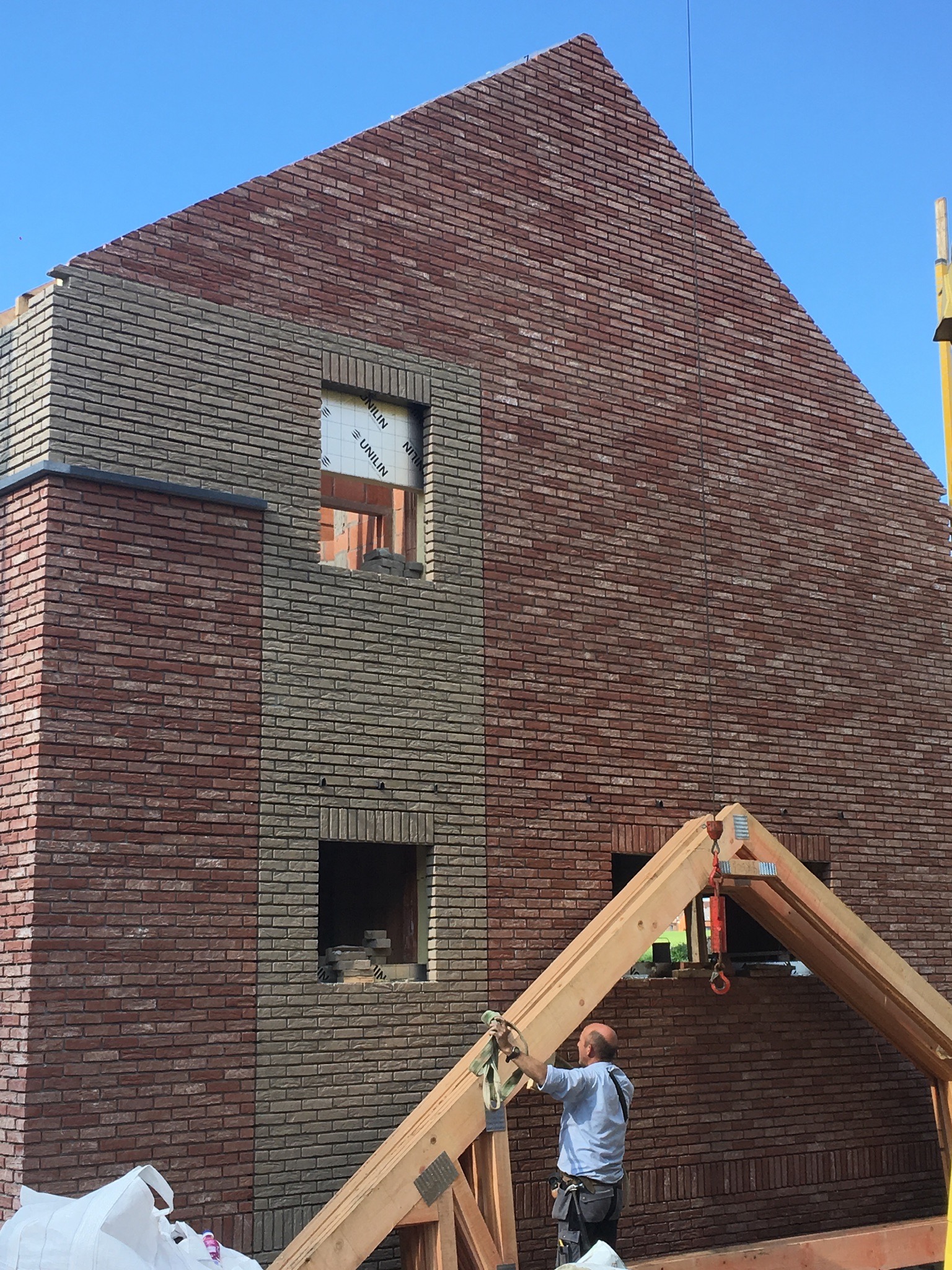 Wznoszenie murowanej ściany domu z cegły klinkierowej w dwóch odcieniach, z widocznymi otworami okiennymi i drewnianą konstrukcją dachu przygotowaną do montażu, pracownik przytrzymuje konstrukcję.