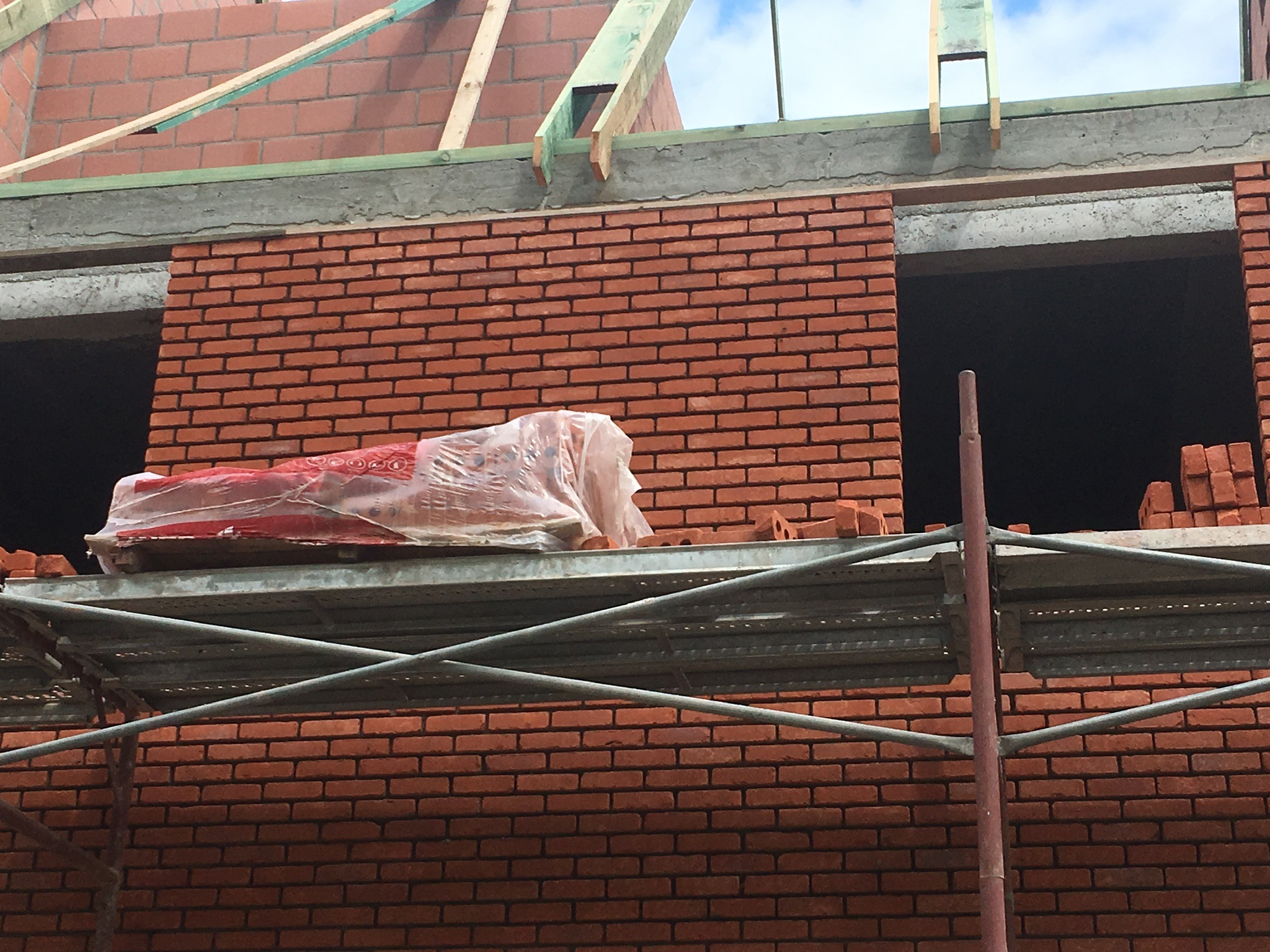 Czerwona ściana z cegły w trakcie budowy, widoczne otwory okienne, rusztowanie z materiałem budowlanym w folii, drewniane elementy konstrukcyjne dachu.