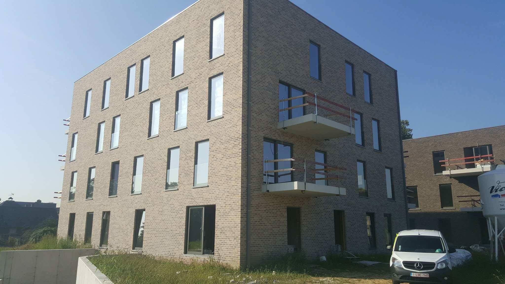 Nowoczesny budynek mieszkalny z jasnej cegły z balkonami z drewnianymi barierkami i dużymi oknami, widoczny samochód dostawczy z przodu.