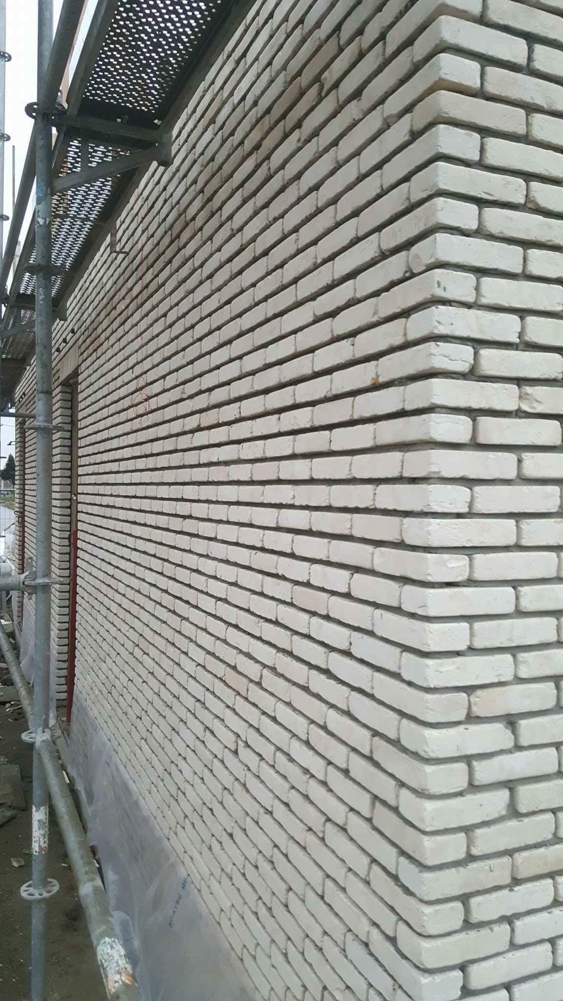 Ściana budynku w trakcie murowania z jasnej cegły, widoczne rusztowanie i folia ochronna u podstawy ściany.