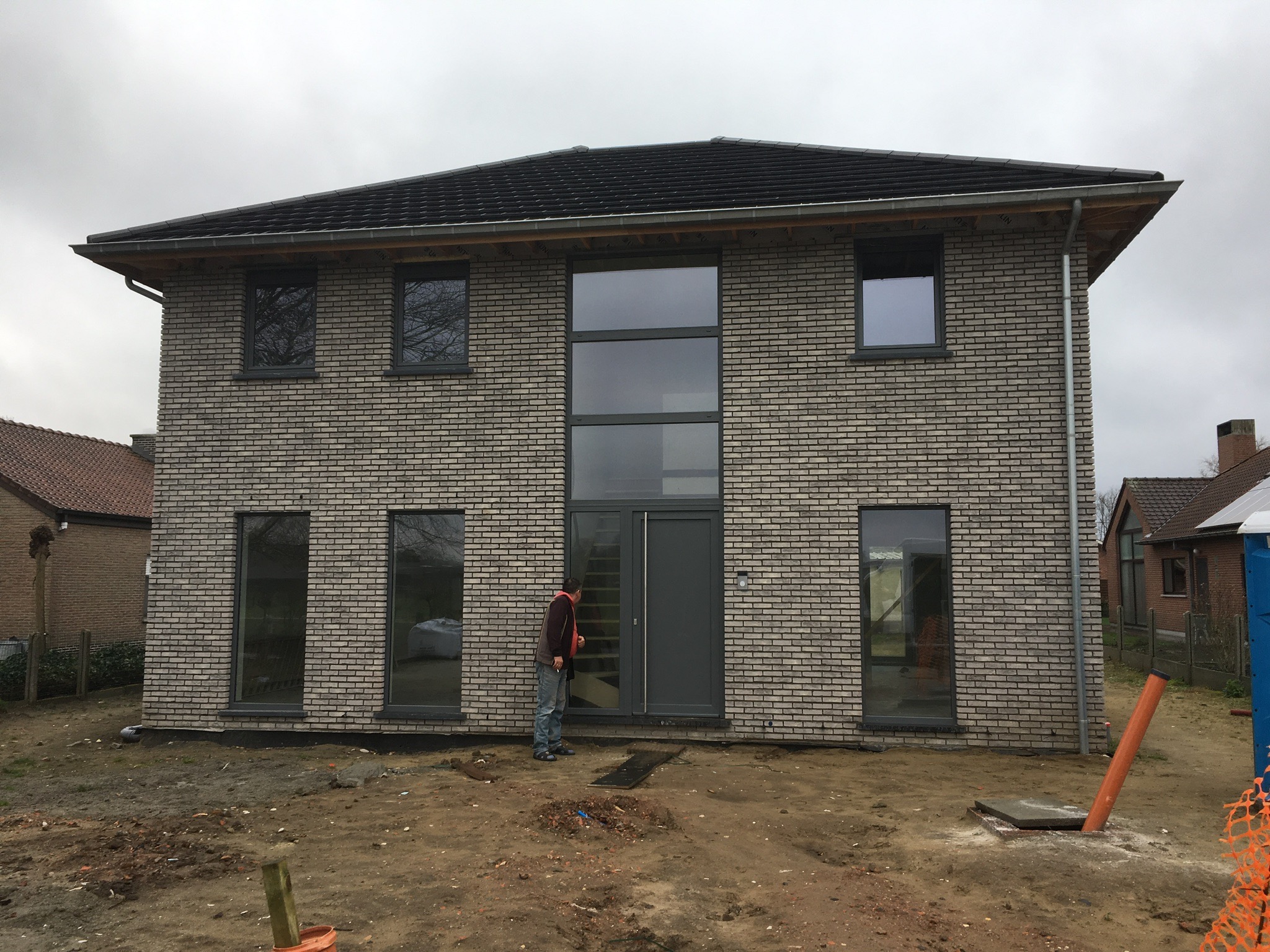Dwukondygnacyjny dom z szarej cegły z dużymi oknami i ciemnoszarą stolarką, widoczny mężczyzna stojący przed wejściem, teren wokół w trakcie zagospodarowania.