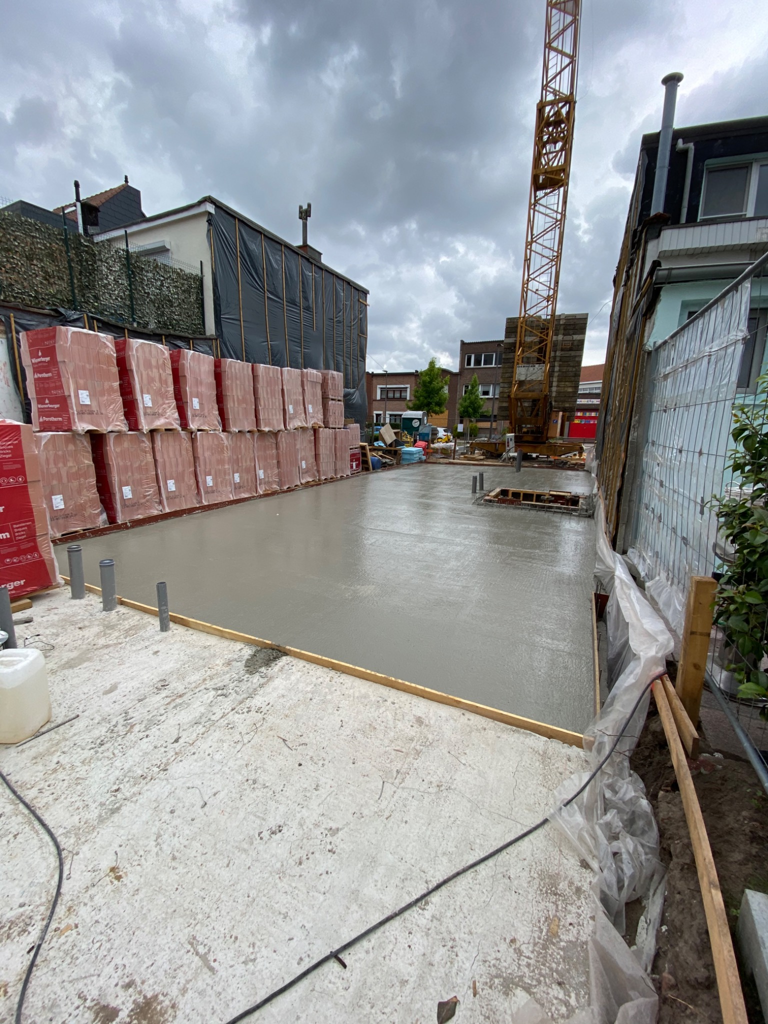 Świeżo wylany fundament z widocznymi przewodami, szalunkiem i paletami cegieł w tle, na budowie z dźwigiem w pochmurny dzień.