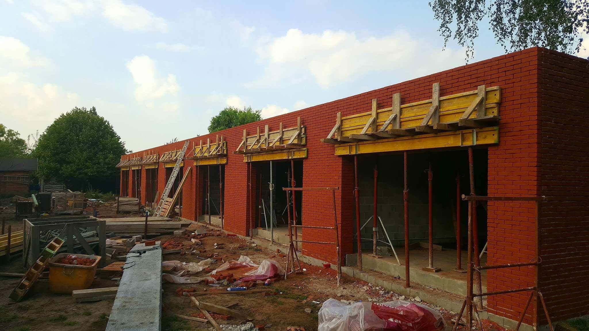 Budowa parterowego budynku z czerwonej cegły z drewnianymi szalunkami nad otworami okiennymi, widoczne rusztowania i materiały budowlane na placu budowy.
