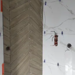 Układanie płytek imitujących drewno w jodełkę na ścianie, widoczne krzyżyki dystansowe i poziomica laserowa, obok ściana z białych płytek imitujących marmur z niepodłączonymi gniazdkami...