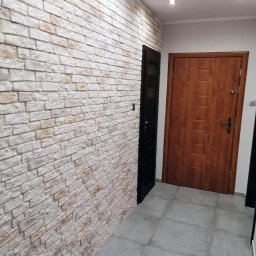 Esewi Bud - Korytarz z dekoracyjną ścianą z cegły w odcieniach bieli i beżu, szarą podłogą z płytek, ciemnymi drzwiami po lewej i drewnianymi drzwiami po prawej stronie.