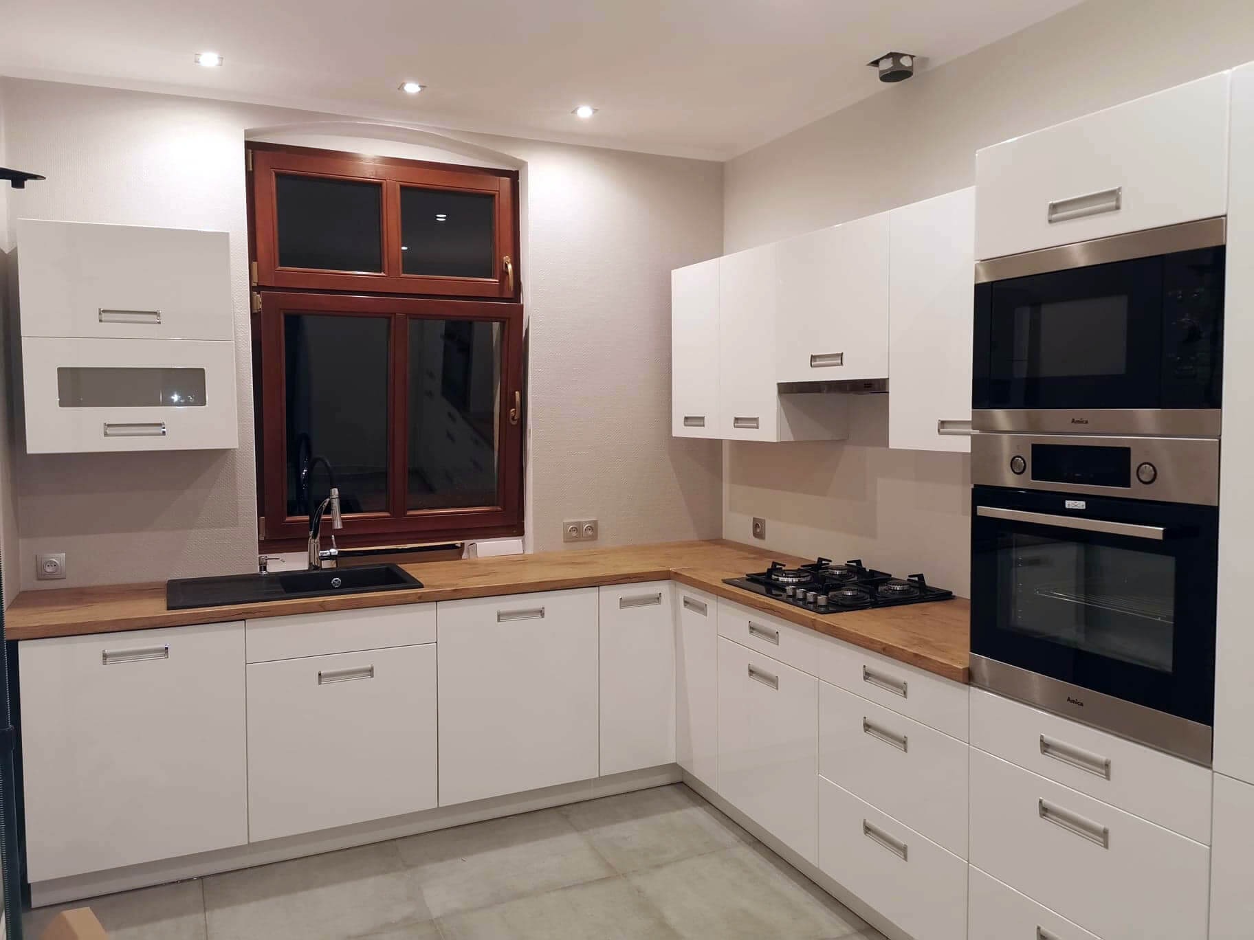 Nowoczesna, jasna kuchnia z białymi szafkami z poziomymi uchwytami i drewnianym blatem, czarny zlew pod oknem z brązową ramą, zabudowany piekarnik i płyta gazowa.