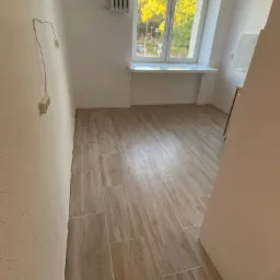 Jasne, podłużne płytki imitujące drewno ułożone na podłodze w pokoju z grzejnikiem pod oknem i wystającą ze ściany instalacją elektryczną.