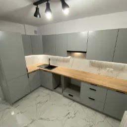 Nowoczesna kuchnia z szarymi szafkami, drewnianym blatem i marmurową podłogą, oświetlona punktowo i taśmą LED pod szafkami wiszącymi.