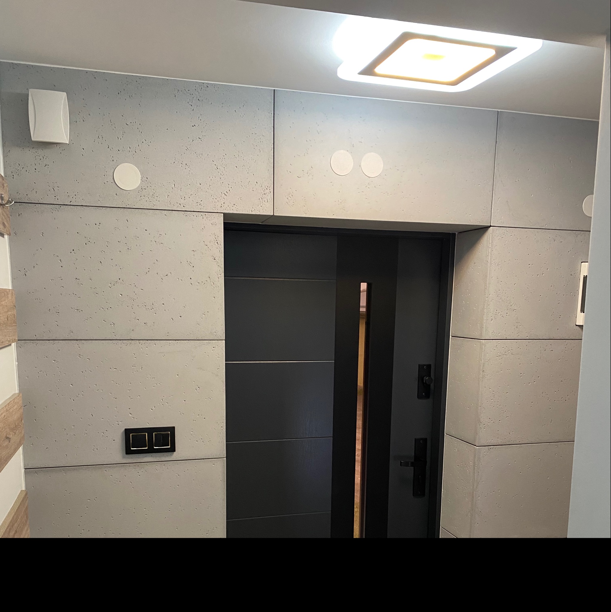 Szara ściana wykończona płytami imitującymi beton, z wkomponowanymi głośnikami, włącznikiem światła i czarnymi drzwiami z pionowym, złotym akcentem, oświetlona kwadratową lampą sufitową.
