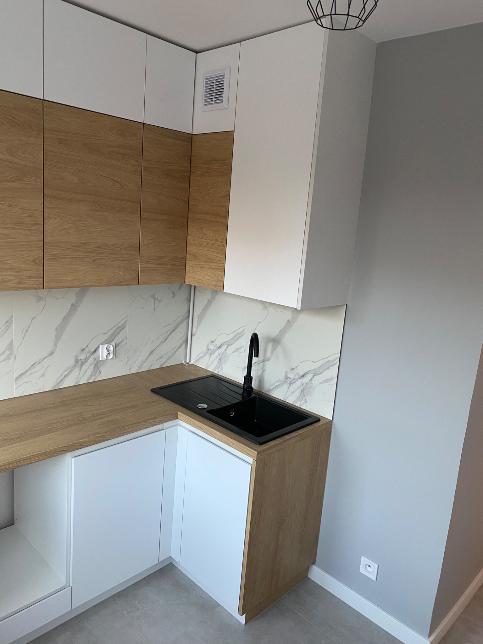 Nowoczesna kuchnia w odcieniach bieli, drewna i szarości, z czarnym zlewem i baterią, marmurowe płytki na ścianie, widoczne gniazdko elektryczne.
