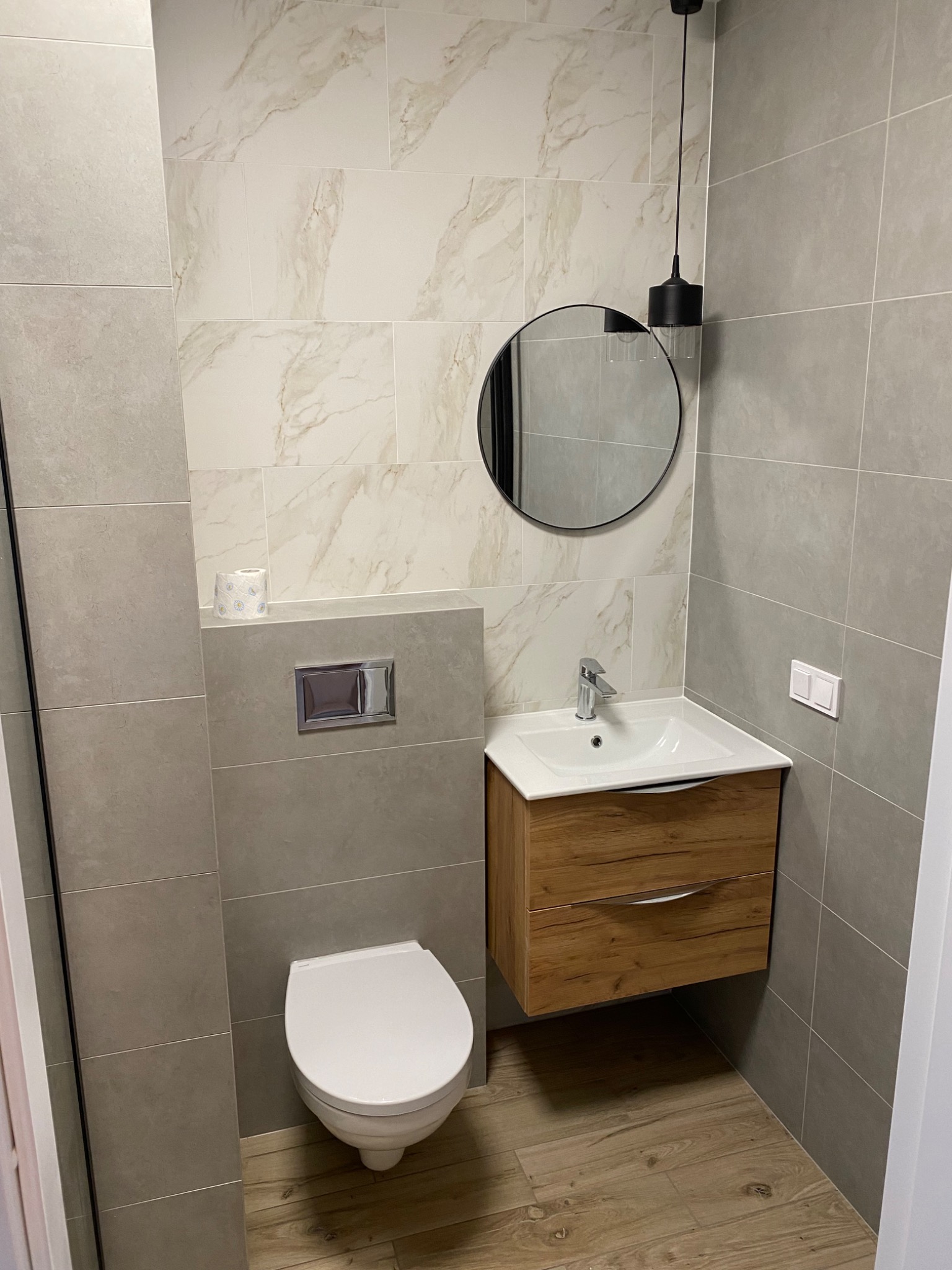 Nowoczesna łazienka z płytkami imitującymi marmur na ścianie, okrągłym lustrem z czarną ramą, wiszącą szafką z umywalką z drewnianym frontem i podwieszaną toaletą.