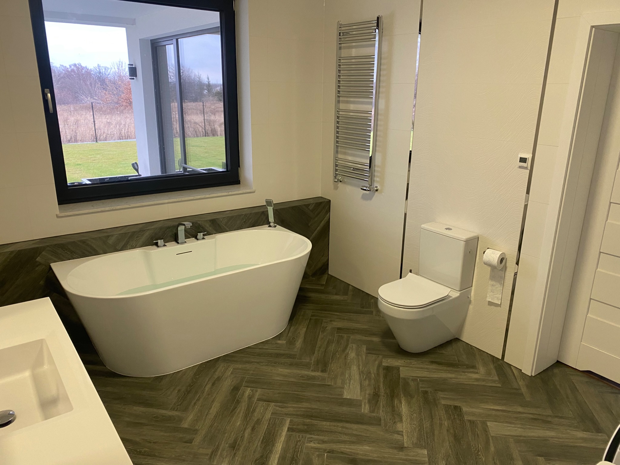 Nowoczesna łazienka z wolnostojącą białą wanną, toaletą typu Geberit i grzejnikiem drabinkowym, wykończona płytkami imitującymi drewno ułożonymi w jodełkę.