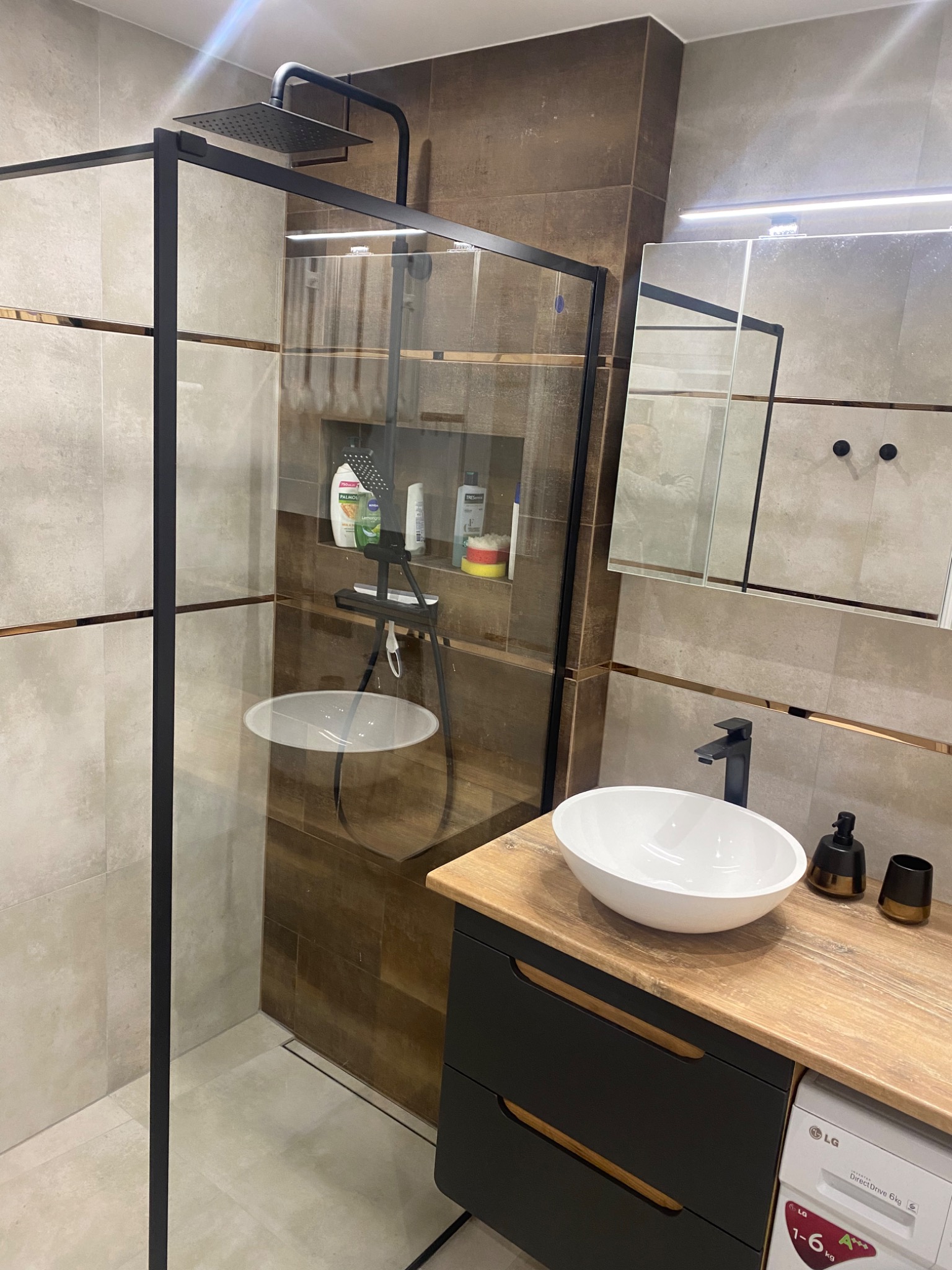 Nowoczesna łazienka z kabiną prysznicową typu walk-in z czarnymi ramami, umywalką nablatową na szafce z drewnianym blatem i pralką LG w zabudowie.
