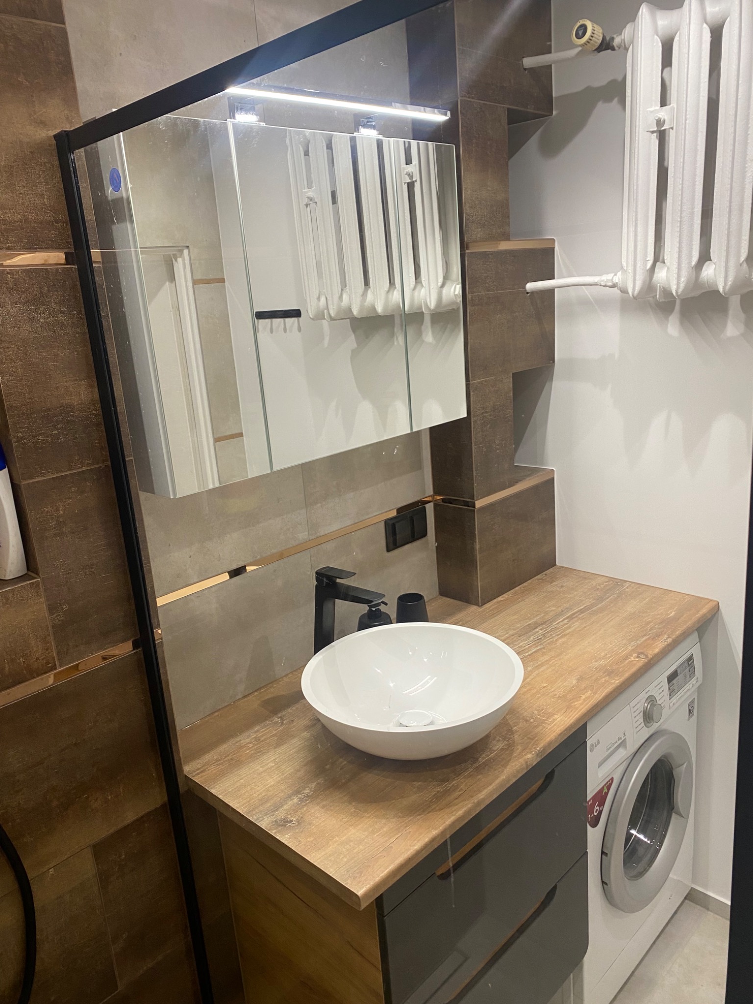 Umywalka nablatowa w nowoczesnej łazience z brązowymi płytkami i drewnianym blatem, pralka LG obok szafki pod umywalką, widoczne odbicie grzejnika w lustrze