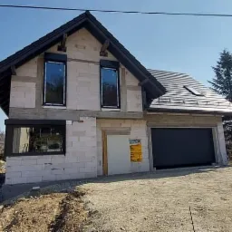 Dom w trakcie budowy z zamontowanymi oknami z roletami zewnętrznymi i bramą garażową, widok z zewnątrz na tle pagórkowatego krajobrazu z kościołem w oddali.