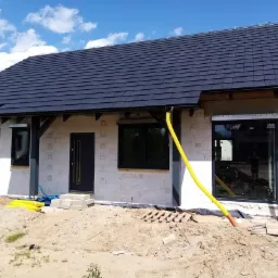 Dom w trakcie budowy z zamontowanymi oknami z czarnymi ramami, zewnętrznymi roletami i ciemnymi drzwiami wejściowymi; widoczne żółte rury odprowadzające wodę z dachu.