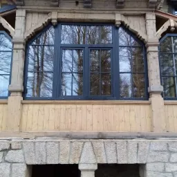 Elewacja drewnianego domu z trzema oknami o nieregularnych kształtach i ciemnych ramach, w których odbijają się drzewa, na kamiennej podmurówce.