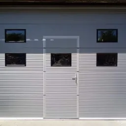 Szara brama garażowa segmentowa z czterema małymi, kwadratowymi okienkami z czarnymi ramami i wbudowanymi drzwiami wejściowymi z klamką.