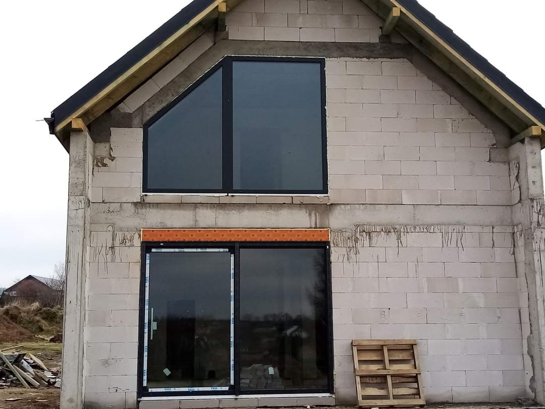 Surowy stan budynku w trakcie montażu stolarki okiennej: okno trójkątne na piętrze i przesuwne drzwi balkonowe na parterze, paleta drewniana oparta o ścianę z betonu komórkowego.