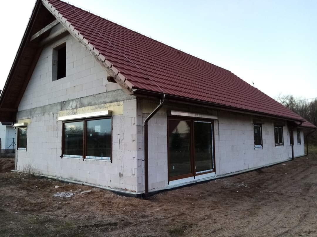 Dom w trakcie budowy z zamontowanymi oknami z brązowymi ramami i częściowo opuszczonymi roletami zewnętrznymi w kolorze białym, widoczne bloczki betonowe i czerwony dach.