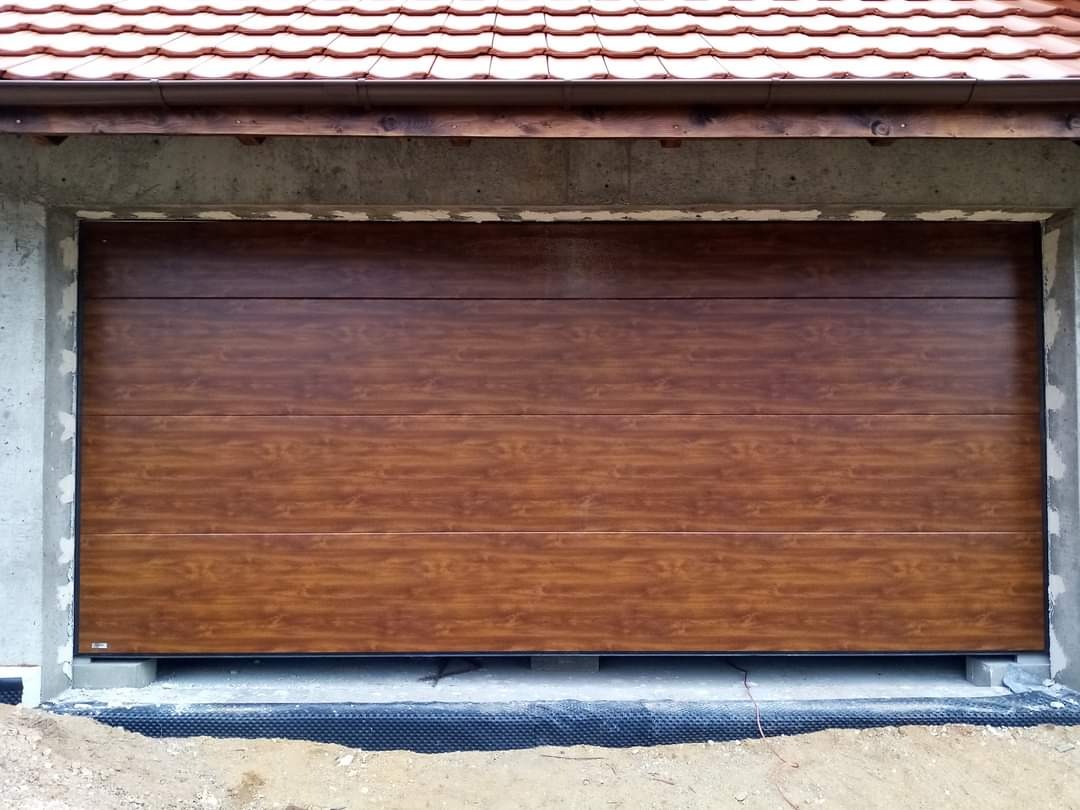 Zamknięta, segmentowa brama garażowa w kolorze ciemnego drewna, zamontowana w surowym, betonowym otworze budynku w trakcie budowy, z widocznym ociepleniem fundamentu pod bramą.