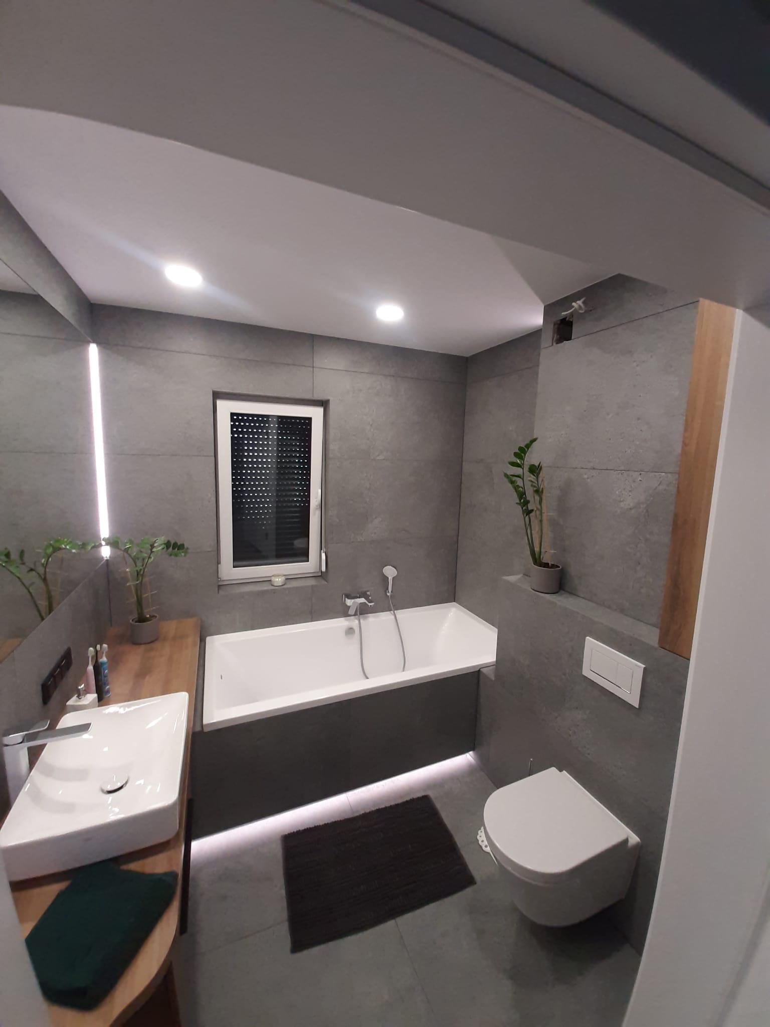 Nowoczesna łazienka z szarymi płytkami, białą armaturą, drewnianym blatem pod umywalką i podświetleniem LED pod wanną.