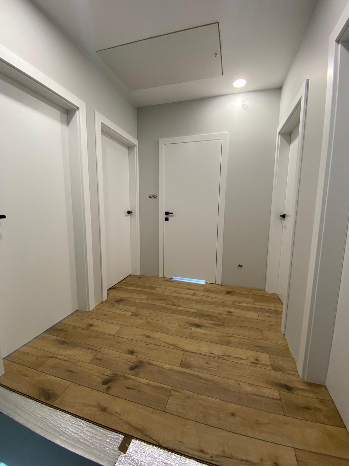Korytarz z trzema białymi drzwiami i panelami podłogowymi w odcieniach brązu, widoczna listwa przypodłogowa i otwór rewizyjny w suficie.