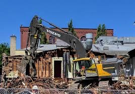 Żółto-szara koparka Volvo w trakcie wyburzania drewnianego budynku z odsłoniętą konstrukcją szkieletową i sterczącymi fragmentami desek, na tle ceglanego budynku i błękitnego nieba.