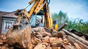 Żółta koparka z łyżką zanurzona w gruzowisku cegieł i fragmentów betonu, w tle fragment dachu domu i zieleń drzew.