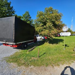 Piotr Czyżowski Transfach - Ciężarówka z czarną plandeką zaparkowana na żwirowym parkingu, w tle zieleń i znak informacyjny. Widoczny fragment tablicy rejestracyjnej.