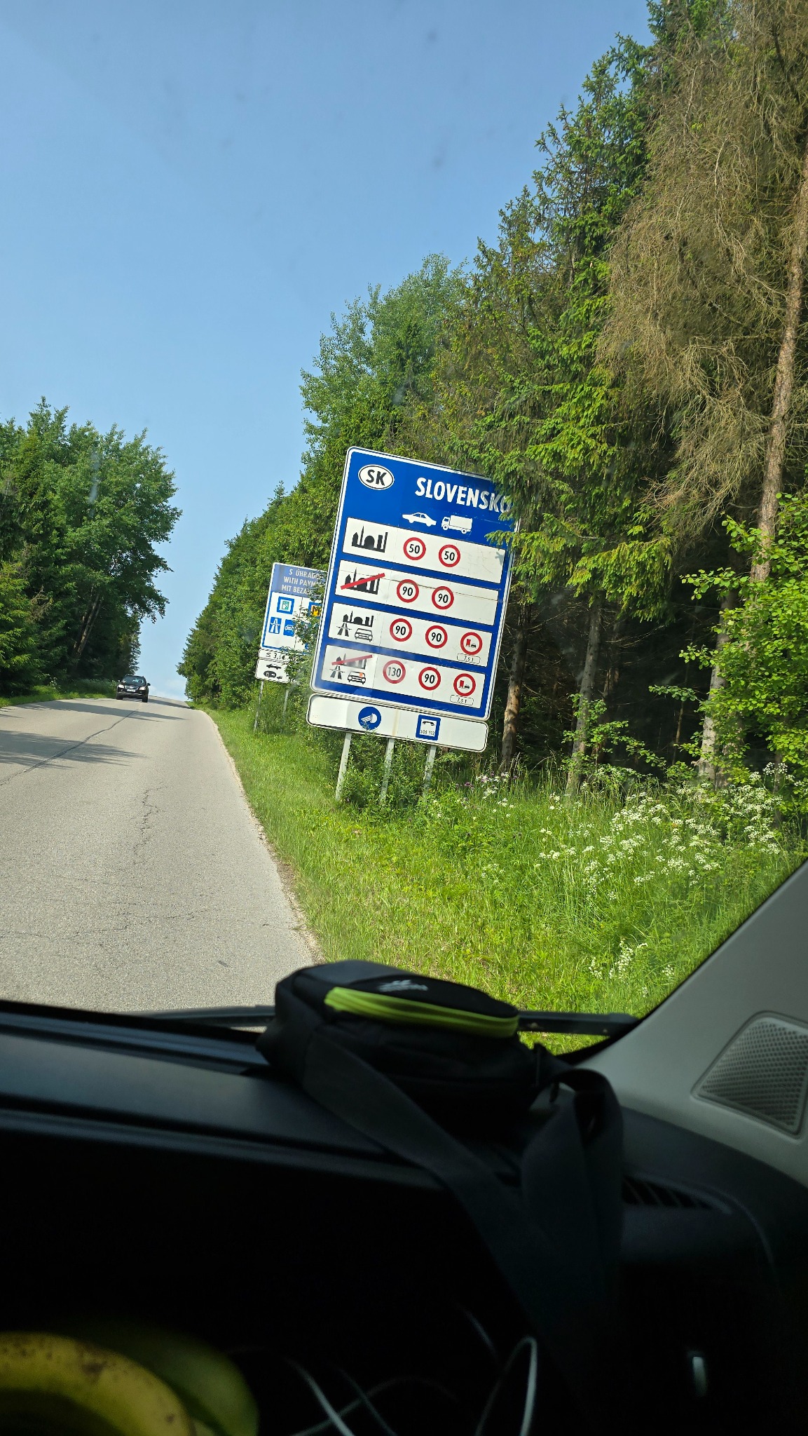 Znak graniczny Słowacji widziany z wnętrza samochodu, z ograniczeniami prędkości dla różnych pojazdów. W tle droga, drzewa i niebieskie niebo.