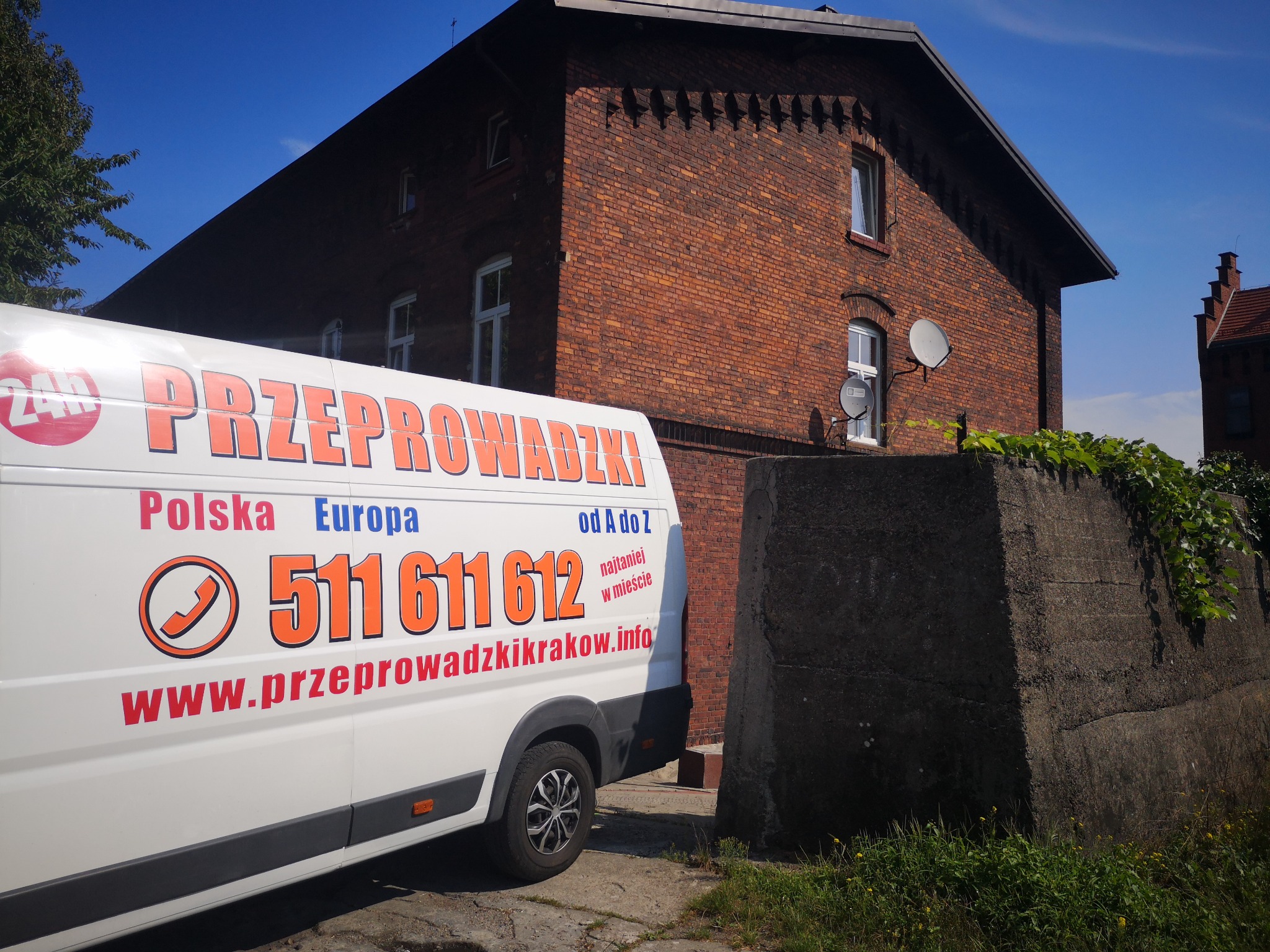Biały bus z pomarańczowym napisem 'Przeprowadzki Polska Europa 511 611 612' zaparkowany przed ceglanym budynkiem z antenami satelitarnymi.